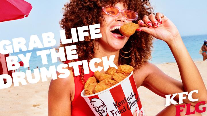 KFC FLG — KFC Happy young woman on the beach holding a buckt of KFC chicken and about to eat a piece. White copy superimposed over image: Grab Life by the Drumstick