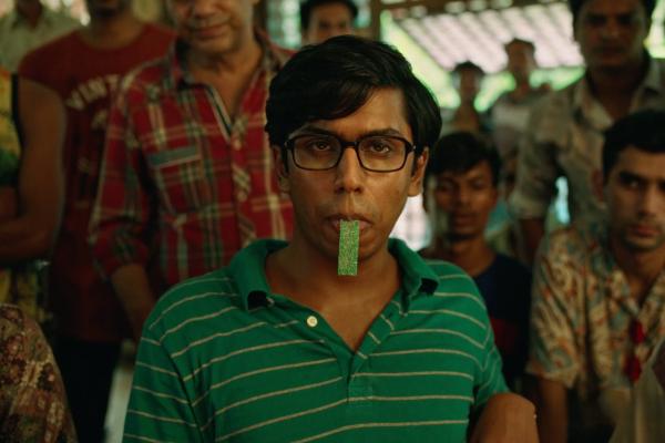 Carrom Board — Chupa Chups A young man with dark hair and glasses wears a green and white striped polo shirt, looking directly at the viewer with a green, rectangular object held between his lips. Several other people are blurred in the background.