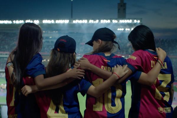 The Bimbo Dream — BImbo and FC Barcelona Four young girls, seen from behind, stand with their arms around each other's shoulders, watching a soccer game in a stadium at night. They are wearing red and blue soccer jerseys with numbers and names on the back. Two of the girls are wearing baseball caps. The stadium lights illuminate the field and the crowd in the background, creating a vibrant atmosphere.