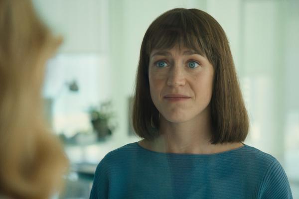 Let's Create Smarter Business — IBM A woman with shoulder-length brown hair cut in a bob with bangs and striking blue eyes looks directly forward with a slightly concerned or serious expression. She is wearing a ribbed teal or blue top. Her gaze is focused, and her mouth is slightly open as if about to speak or having just spoken. The background is blurred, showing a bright, indoor environment, possibly an office, with light-colored walls and reflections.