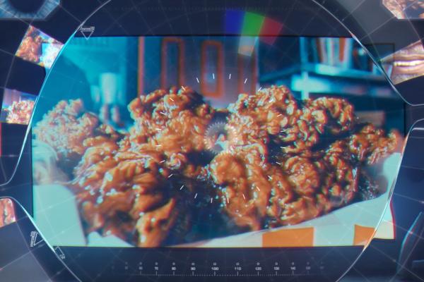 KAIIA — KFC A close-up shot of several pieces of crispy, golden-brown fried chicken in a paper-lined basket. The image is viewed through a stylized, dark, circular frame with a central glowing gear-like icon and radiating white lines, resembling a futuristic lens or display. The background shows hints of a kitchen or restaurant setting.