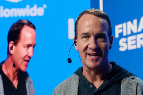 Motivatin' Manning / So Much more -- Nationwide A close-up shot of Peyton Manning, a light-skinned man with short brown hair, smiling and speaking into a headset microphone. He is wearing a grey blazer over a dark hooded sweatshirt. His face is well-lit, and his eyes are looking slightly to the right. In the blurred background, a large blue screen shows the word "Nationwide" on the left and partially visible text "FINA SER" on the right, along with a reflection of Manning's image.