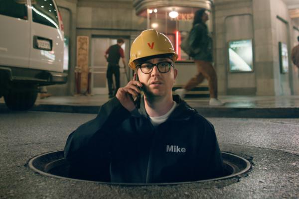 Verizon Now — Verizon Man in a yellow safety helmet on a phone while emerging out of a manhole
