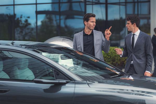 This is Audi — Audi Two young men standing behind an Audi car. They are both wearing suits. The man on the left is point his finger at the man on the right.
