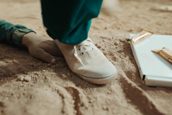 Cadbury 5 Star x Squid Game White sneaker stepping down on stand, on the ground next to the foot is a white piece of debris that has two gold Cadbury 5-Star bars on it