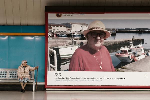 JCDecaux – Meet Marina Prieto An image of 99 year old Marina Prieto sitting in the Madrid subway station next to a large billboard featuring one of her instagram posts from her account "marinaprieto1932"