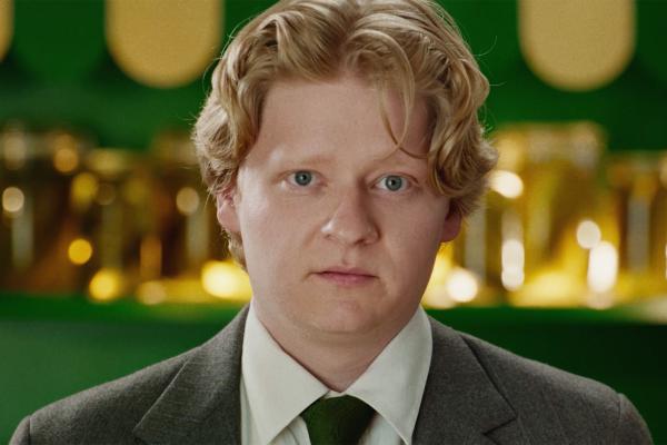 Make it Legit — H&R Block A fair-skinned man with blond, wavy hair and blue eyes looks directly at the camera with a neutral expression. He is wearing a grey suit, white shirt, and green tie, standing in front of a green and gold background.