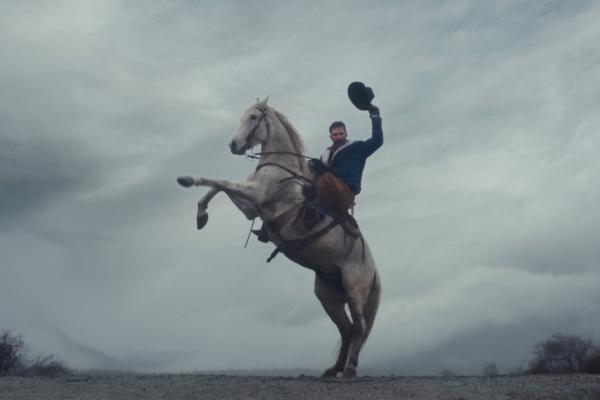 Bring Out Your Adventurous Side — Mitsubishi A man with a mustache, wearing a blue jacket and brown chaps, rides a white horse that is rearing up on its hind legs. The man is raising his hat in the air, appearing celebratory, against a backdrop of a cloudy, grey sky and a barren landscape.
