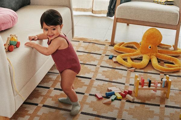 Supporting First Steps — Al-Futtaim IKEA A baby stands with the help of a couch. On the floor are kids toys -- wooden blocks and a stuffed animal octopus