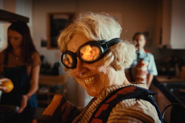 More For Everything You Love — Bimbo A close-up shot shows an older woman with blonde hair, wearing black goggles that reflect a fiery orange glow, smiling widely with her teeth showing. She is wearing a patterned shirt with a dark blue and red design. In the blurred background, another woman in a dark apron holds a yellow fruit, and a man in a light blue shirt and red apron stands further back.