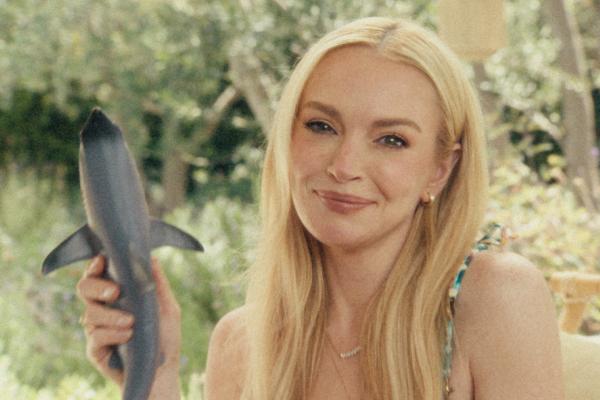 Total Glow Up / Different — Verizon A smiling Lindsay Lohan, with long blonde hair, holds up a small, dark grey shark toy. She wears a patterned top and a delicate necklace. The background is blurred with green foliage.