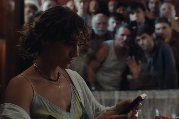 It's Not a Place — Ministry of Equality of Spain A young woman with dark, curly hair looks down at a phone in her hands, wearing a light-colored top and a cardigan, with a cross necklace visible. Behind her, a blurred crowd of people, mostly men, are visible, some looking towards her.