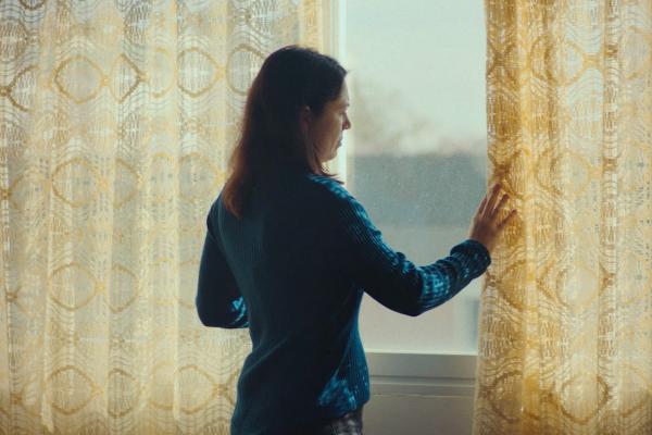 Not One More, Not One Less — Ministry of Equality Woman at a window placing her hand on the curtain