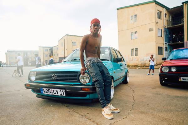 Forever Golf — Volkswagen A young, shirtless man with a red head covering and dark pants leans casually on the hood of a teal vintage Volkswagen Golf. He is looking directly at the camera. The car has a German license plate that reads "WOB CV 17". In the background, there are apartment buildings and other people, including another red vintage car on the right. The setting appears to be an urban outdoor area with a light sky.