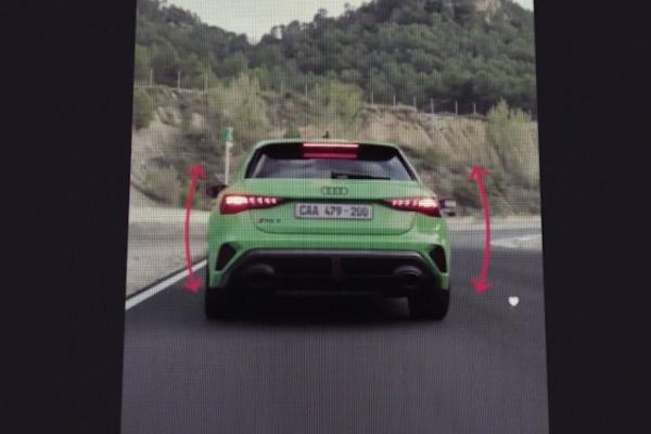 Tiktok Test Drive — Audi A close-up of a smartphone screen displaying the rear view of a bright green Audi RS3 hatchback on a race track. Red curved arrows on either side of the car suggest interactive controls or a visual representation of movement. The car's license plate reads "CAA 479-200". In the background, green trees and hills are visible under a light sky.