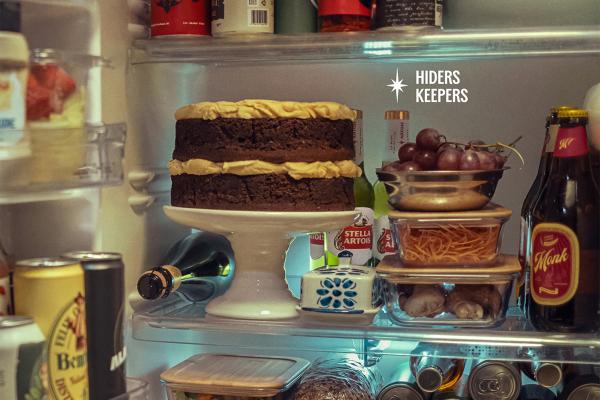 Shot of the inside of a refrigerator. There are assorted items, including a large chocolate cake, spaghetti in a glass container. Hidden behind the items are a few bottles of Stella Artois beer. There is text on the image that says "Hiders Keepers"